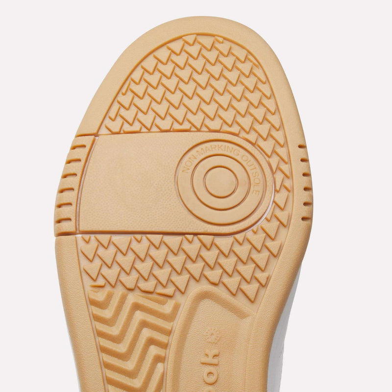 Close-up of a light brown shoe sole featuring various tread patterns, such as zigzags and crosshatch designs, with the text NON-MARKING OUTSOLE displayed inside a circle near the center. This detail emphasizes the quality craftsmanship of the Reebok Men’s Court LXS Shoes—a premium men’s court shoe built with full grain leather for superior durability and style.