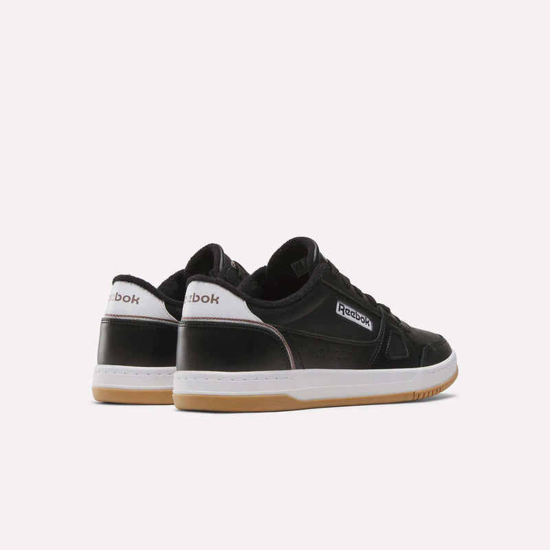A pair of Men’s Court LXS Shoes by Reebok, featuring a black full grain leather upper, white soles, and brown gum bottoms, is displayed from the back and side against a plain white background.