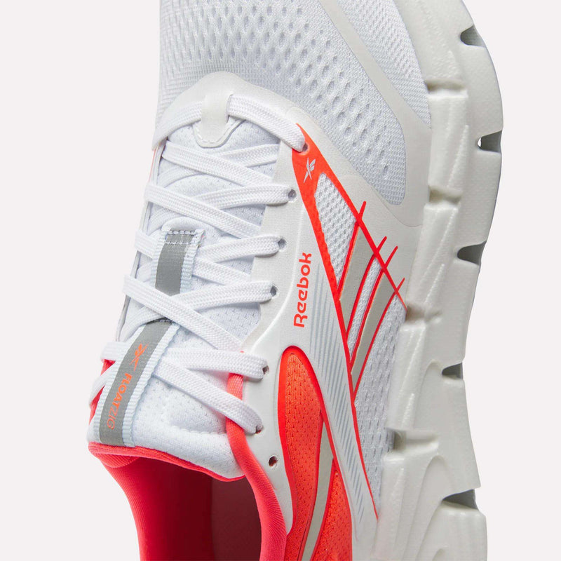 Close-up of a white Reebok Women's FloatZig 2 Running Shoe, featuring orange and red accents, a breathable mesh upper, white laces, and a thick, textured white sole with Superfloat+ cushioning for springy comfort. The Reebok logo is visible on the side near the laces.