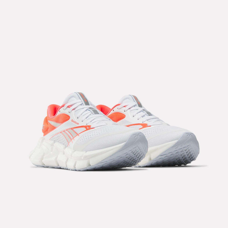 A pair of Reebok Women's FloatZig 2 Running Shoes in white, featuring light gray soles and orange accents, is displayed against a plain white background. These sneakers boast a modern design with prominent Reebok branding on the sides and tongue, and provide enhanced comfort with Superfloat+ cushioning for a responsive running experience.
