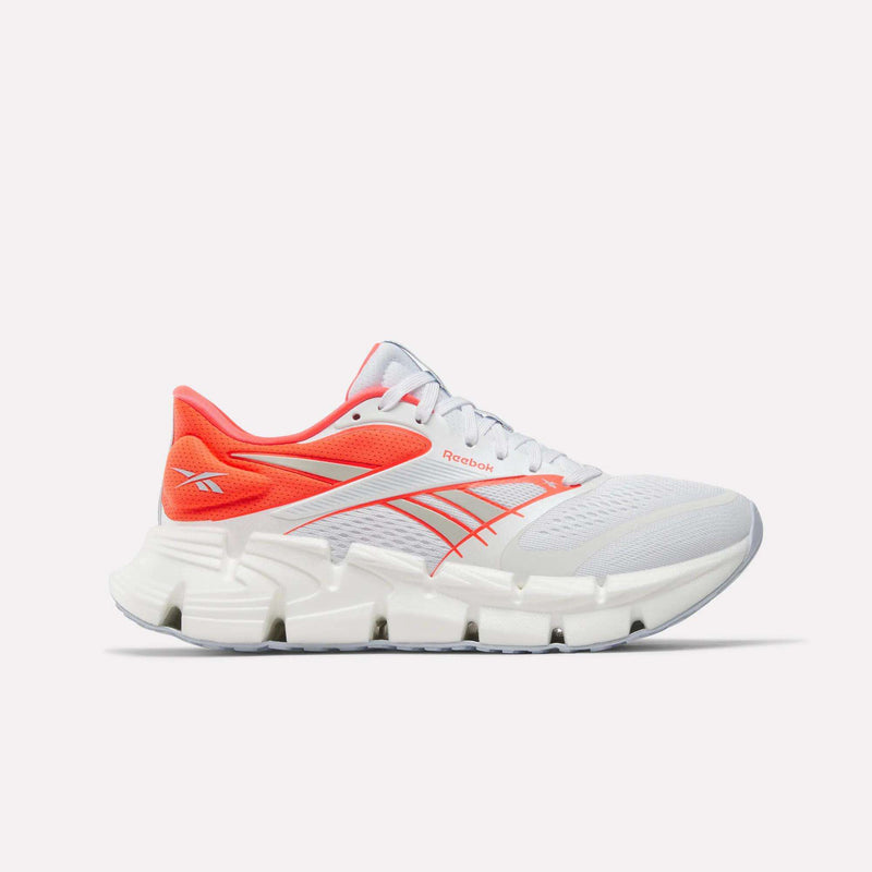 A pair of Women's FloatZig 2 Running Shoes by Reebok in white, highlighted with vibrant orange accents on the heel and details, showcases the signature Reebok logo. The shoes feature a thick, segmented white sole designed for enhanced Superfloat+ cushioning, providing springy comfort with every step. Displayed against a plain white background.