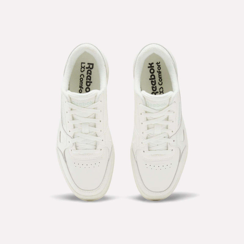 Two white Reebok Women’s Court LXS Shoes are viewed from above on a plain, light background. Reebok and LJ Comfort are visible on the insoles in black text. These women’s court shoes feature full grain leather, white laces, and a classic, simple design.