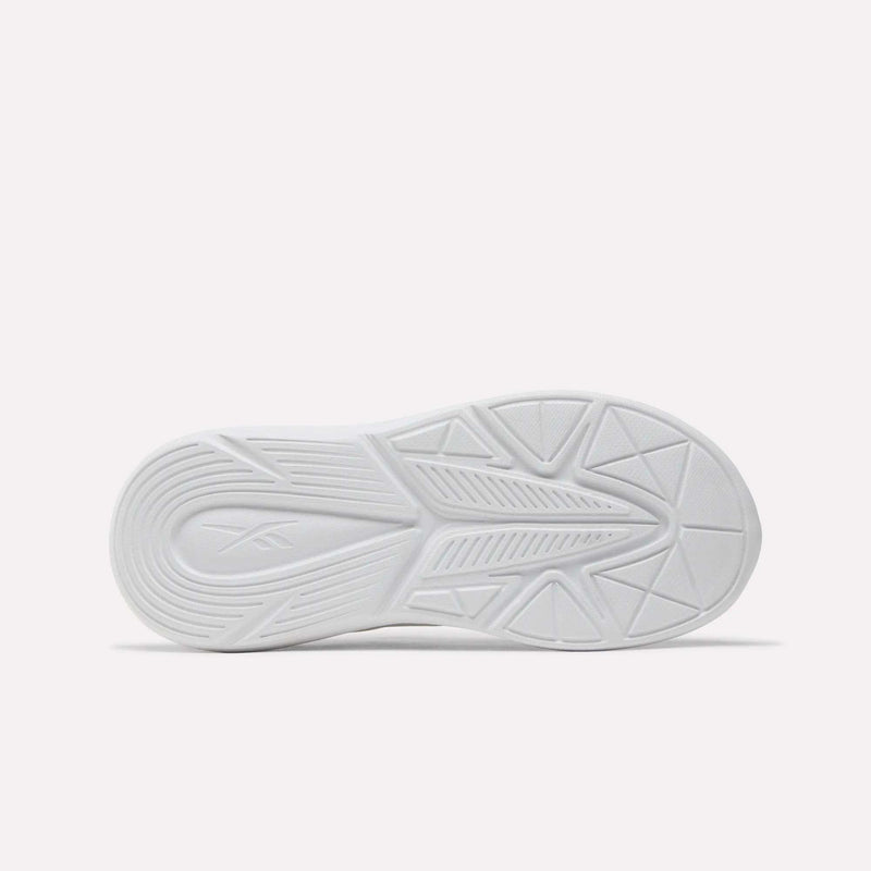 The image shows the bottom view of a white Reebok Rush Runner 6.0 Elastic Lace Top Strap Running Shoe for little kids, featuring geometric tread patterns and an embossed Reebok logo on the left, plus EVA cushioning for comfort. The background is plain and light-colored.