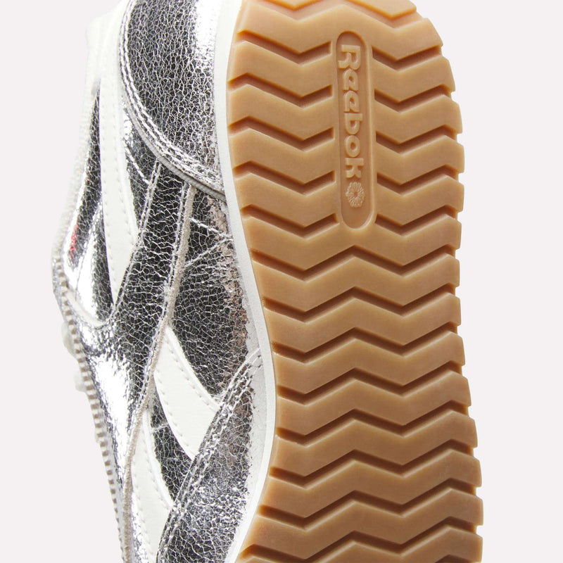 Close-up of the sole of the Women’s Classic AZ Shoes by Reebok, showcasing a tan gum outsole and a metallic silver upper with white stripes, highlighting the enduring style of classic retro runners.