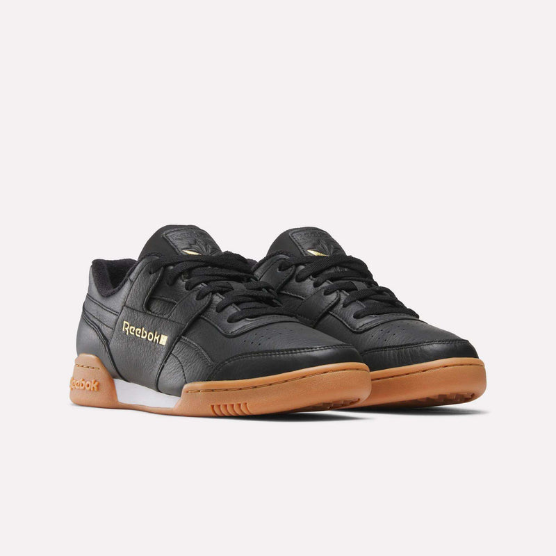A pair of Reebok Workout Plus Shoes in black, featuring gum soles, gold Reebok branding, and black laces, are positioned side by side on a plain white background, showcasing the classic ’80s fitness style.