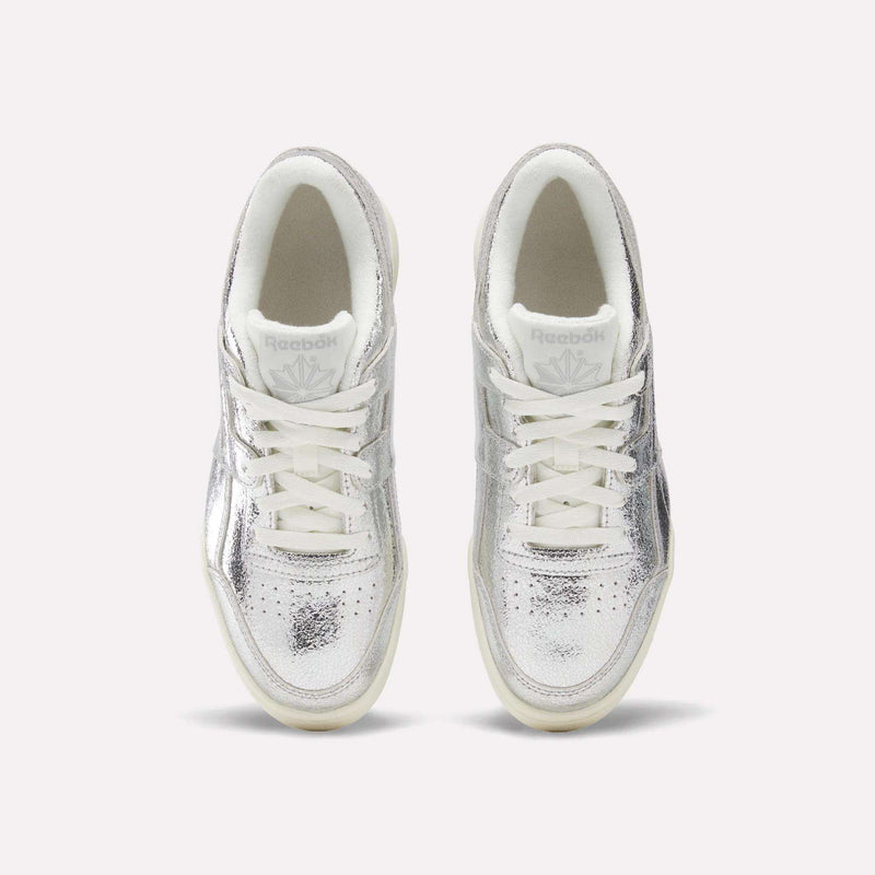 Here’s a rewritten version using the provided product data: A top-down view of Reebok Women’s Workout Plus Shoes crafted in premium garment leather, showcasing their shiny silver finish with white laces and soles, elegantly positioned side by side against a plain light background.