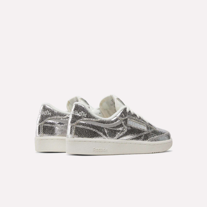 A pair of Reebok Women’s Club C 85 Shoes in metallic silver leather, featuring white soles, are positioned side by side and angled slightly away on a plain light background.