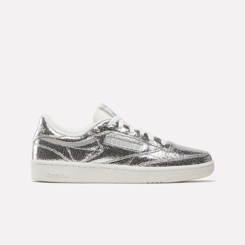 A side view of the Reebok Women’s Club C 85 Shoes in Silver Metallic/Chalk/New Grey 1 shows off their textured metallic leather upper and crisp white sole against a plain light background. Inspired by classic tennis style, this design combines comfort with eye-catching shine.