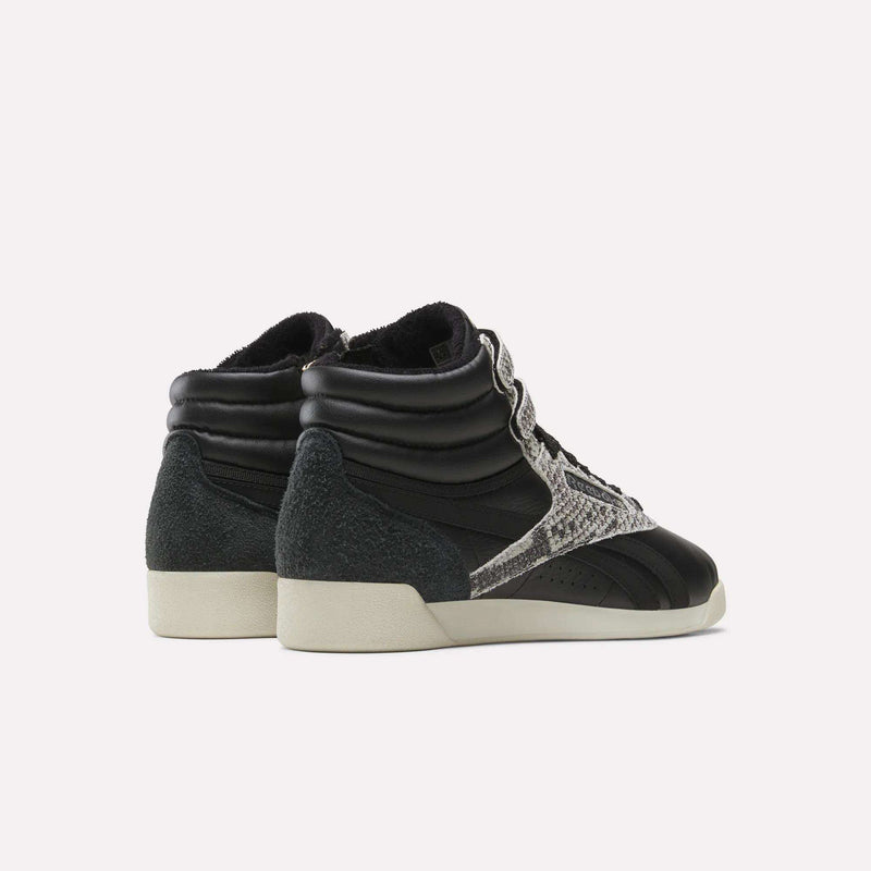 A pair of Reebok Women’s Freestyle Hi Zip Shoes in black, featuring white soles, suede heels, and snakeskin-patterned side stripes inspired by classic retro sneakers, displayed from the back and side against a plain white background.