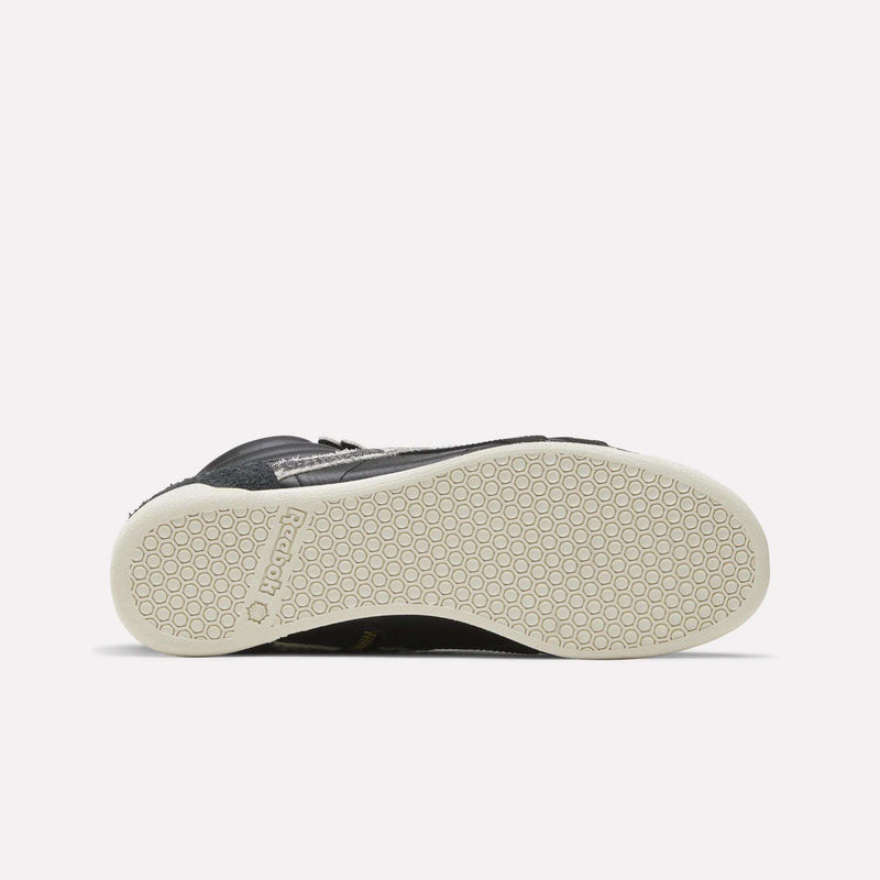The image shows the bottom view of a black Women’s Freestyle Hi Zip Shoe by Reebok with a white sole featuring a hexagonal grip pattern and the Reebok logo near the heel, reminiscent of classic retro sneakers often seen in women’s leather sneaker collections.