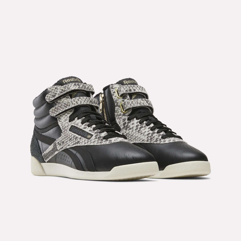 A pair of Reebok Women’s Freestyle Hi Zip Shoes featuring black accents, a grey snakeskin pattern, padded collars, double Velcro straps, and white soles is displayed on a plain white background.