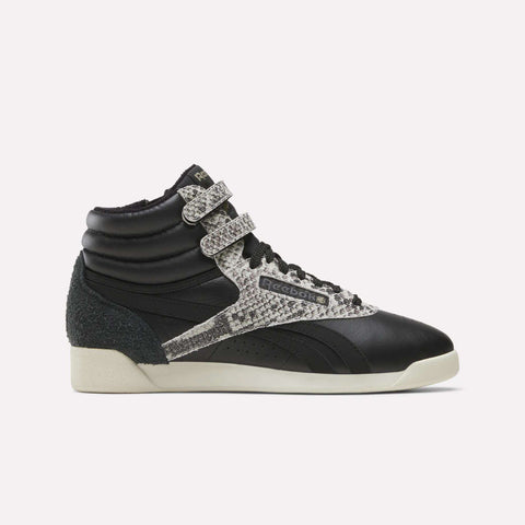 An Almostblack/Vintagechalk/Gold Women’s Freestyle Hi Zip Shoe by Reebok is displayed in side profile against a plain white background, featuring dual velcro straps, snakeskin-patterned accents, black laces, and an off-white sole. These retro-inspired sneakers blend iconic Reebok design with the comfort and durability of women’s leather footwear.