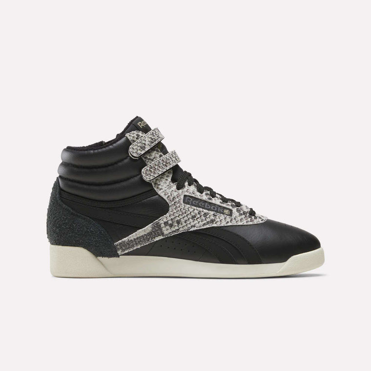 An Almostblack/Vintagechalk/Gold Women’s Freestyle Hi Zip Shoe by Reebok is displayed in side profile against a plain white background, featuring dual velcro straps, snakeskin-patterned accents, black laces, and an off-white sole. These retro-inspired sneakers blend iconic Reebok design with the comfort and durability of women’s leather footwear.
