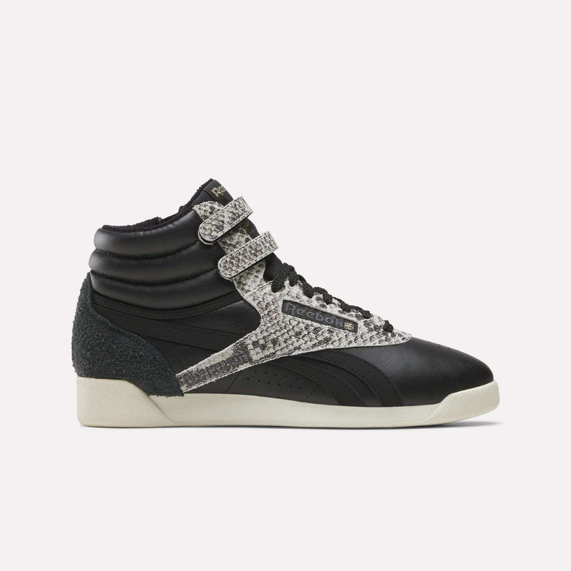 An Almostblack/Vintagechalk/Gold Women’s Freestyle Hi Zip Shoe by Reebok is displayed in side profile against a plain white background, featuring dual velcro straps, snakeskin-patterned accents, black laces, and an off-white sole. These retro-inspired sneakers blend iconic Reebok design with the comfort and durability of women’s leather footwear.