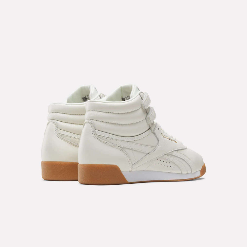 Pair of Reebok Women's Freestyle Hi Shoes in off-white leather with gum-colored soles, featuring Velcro straps and shown from the back and side on a plain white background—perfect for women's fitness routines.