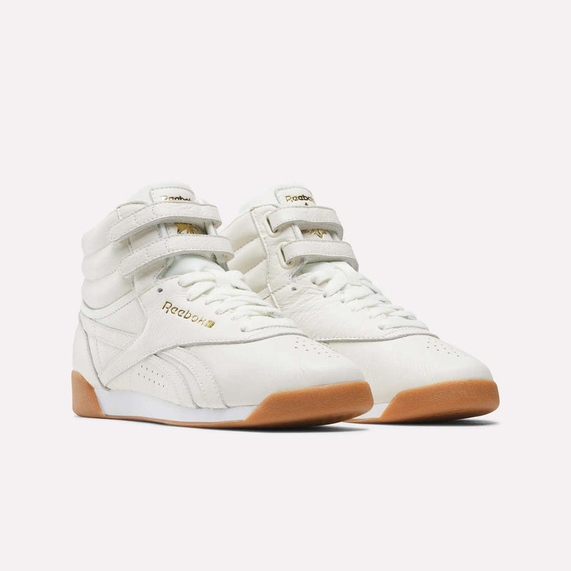 A pair of Reebok Women's Freestyle Hi Shoes in white, featuring gum soles, double velcro straps, and gold Reebok branding, displayed on a plain white background—perfect for elevating any women's fitness wardrobe with both style and support.