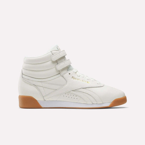The Reebok Women's Freestyle Hi Shoes in Chalk/Gold/Gum feature a classic high-top silhouette with two Velcro straps, white laces, and a light brown gum sole. Shown in side profile against a plain background, these timeless sneakers are ideal for women’s fitness routines or effortless everyday style.