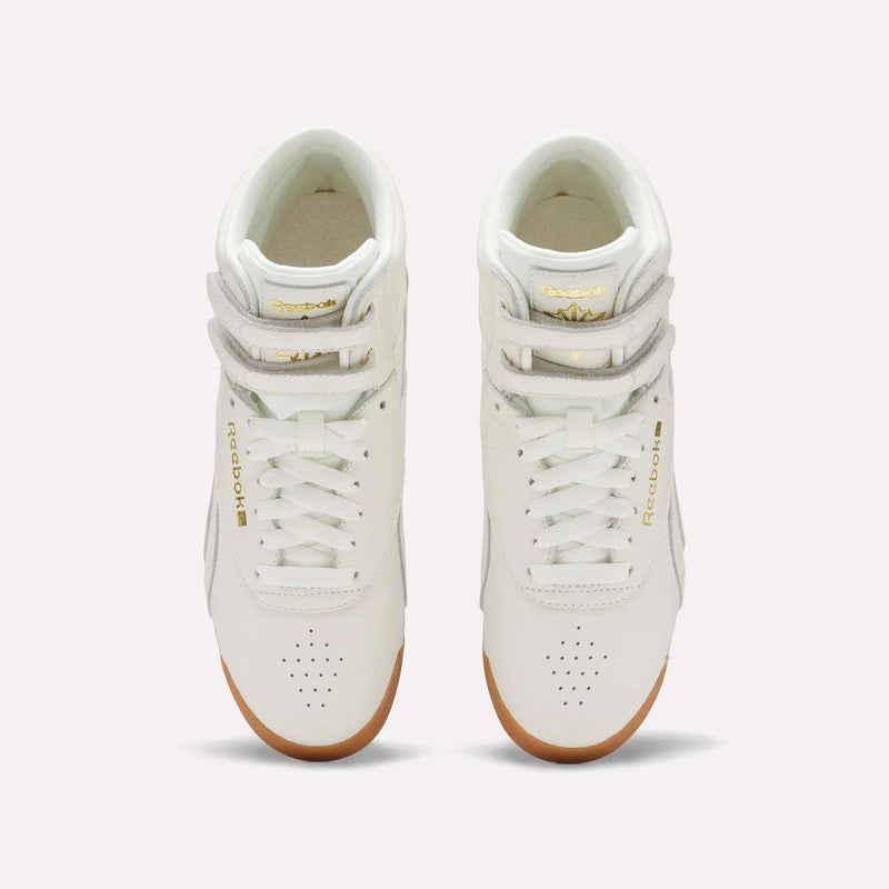 A pair of Reebok Women's Freestyle Hi Shoes in white, featuring gold accents and gum soles, designed for fitness, are displayed from an overhead perspective on a plain light background.