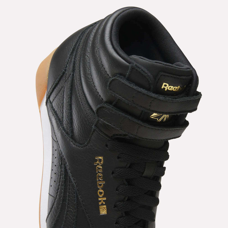 Close-up of a black Reebok Women’s Freestyle Hi Shoes high-top sneaker featuring two ankle straps and gold lettering on the side and tongue.