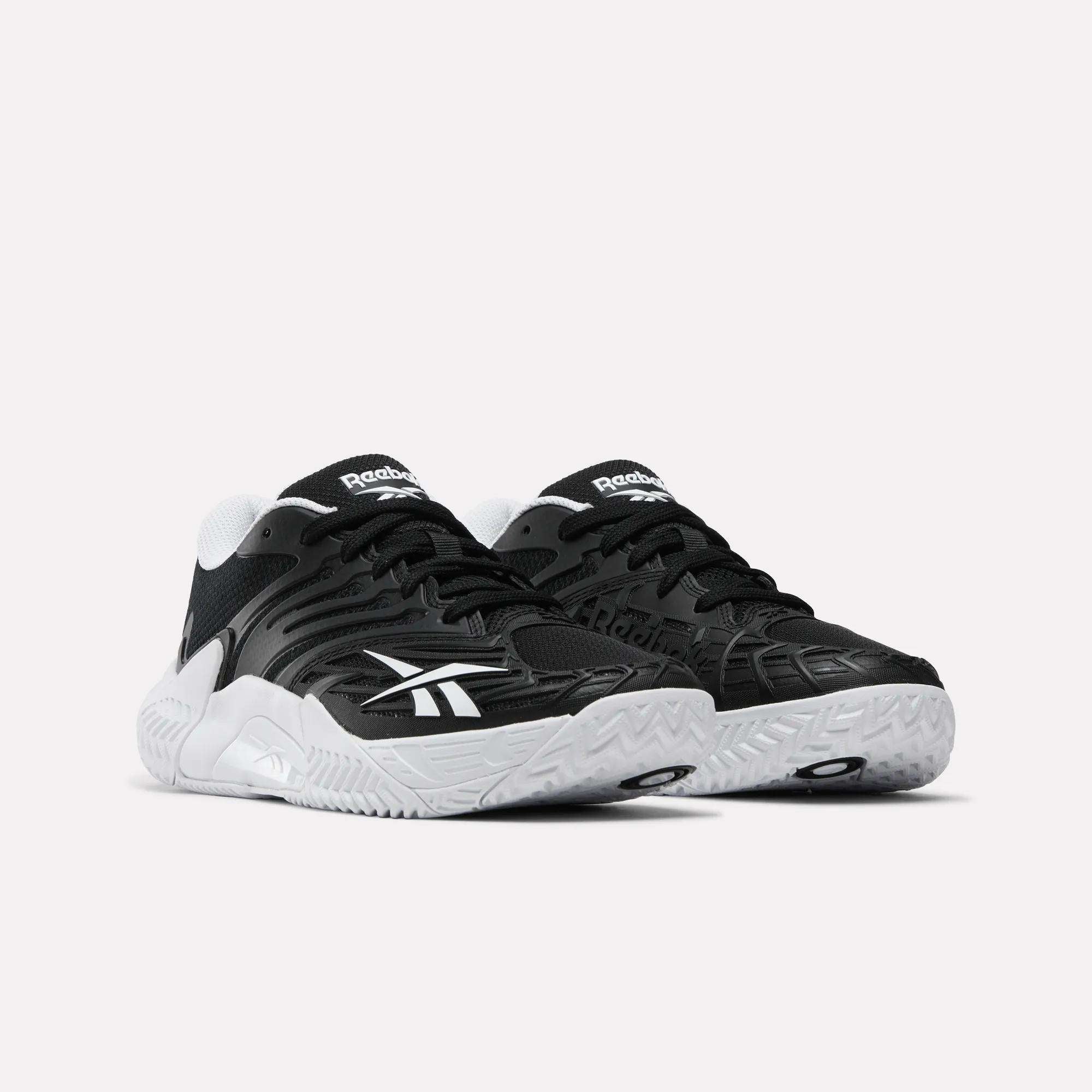 A pair of Reebok Women's Phase Evo Tennis Shoes with black uppers, white logos, white midsoles, and textured soles is displayed side by side on a light background.