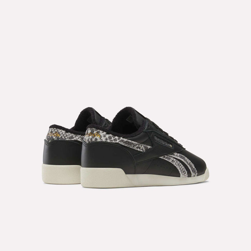 A pair of Reebok Women’s Freestyle Lo Shoes featuring black low-top design, white soles, grey and black snakeskin-patterned accents, and classic logo details is displayed on a plain white background.