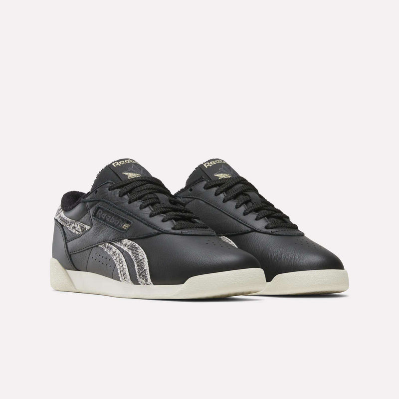 A pair of Reebok Women’s Freestyle Lo Shoes in black, showcasing off-white soles and gray snakeskin-patterned accents on the sides, are displayed against a plain white background.