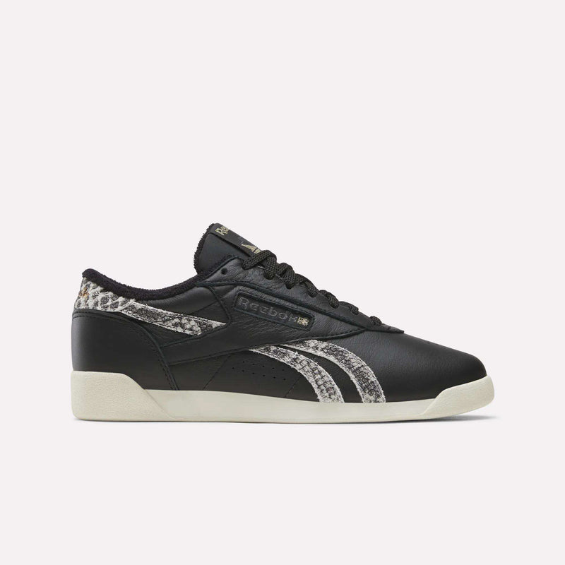 Almostblack Reebok Women’s Freestyle Lo Shoes featuring Vintagechalk soles and Reeboklightgold snakeskin-patterned accents on the side stripes and heel, displayed in a left side view against a plain white background.