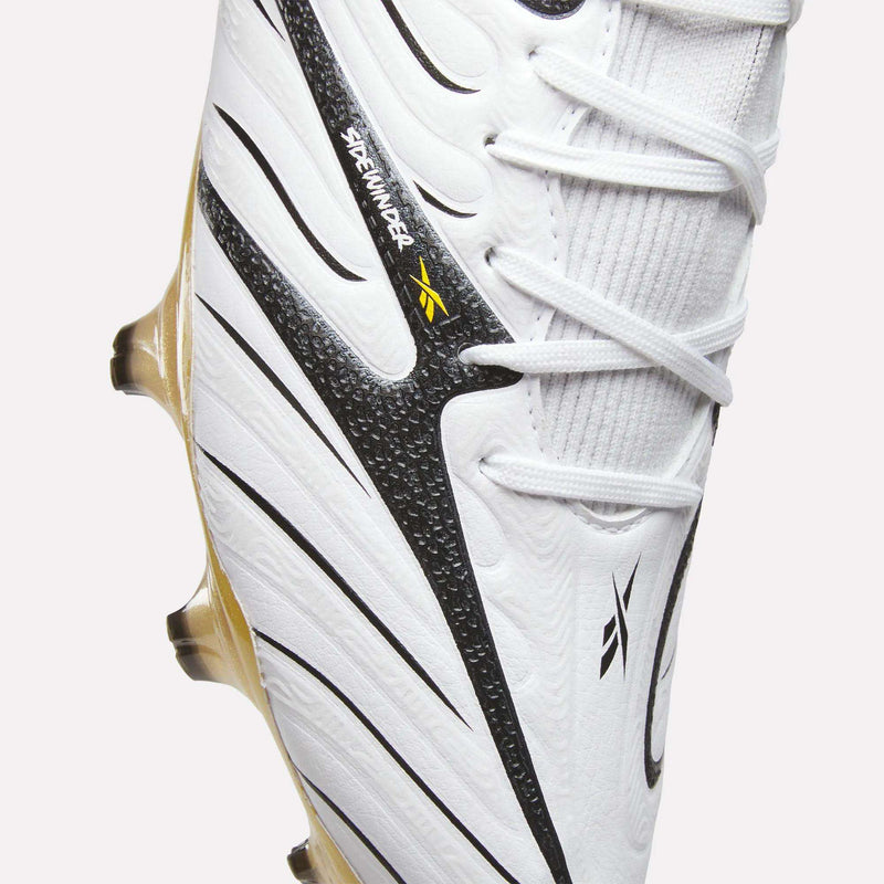 Close-up of the Reebok Sidewinder 26 Elite Firm Ground Cleats in white, featuring black and gold accents, textured detailing for improved ball control, and Reebok branding near the laces—ideal for play on firm grass surfaces.
