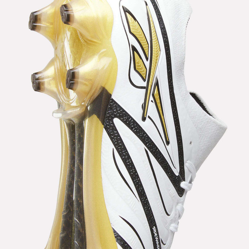 Close-up of the Reebok Sidewinder 26 Elite Firm Ground Cleats in white and gold, featuring three gold studs and a dynamic black and gold line design on the upper and sole for enhanced ball control.