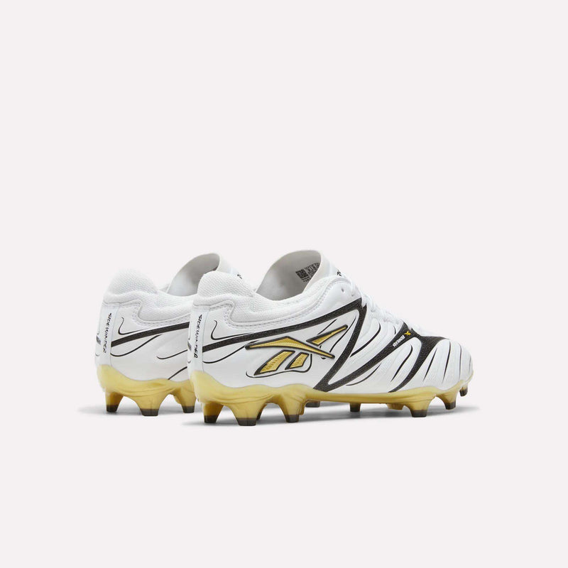 A pair of Reebok Sidewinder 26 Pro Firm Ground Cleats in white, featuring black and gold accents, including a gold Reebok logo and gradient gold soles, designed to enhance ball control, displayed against a plain white background.