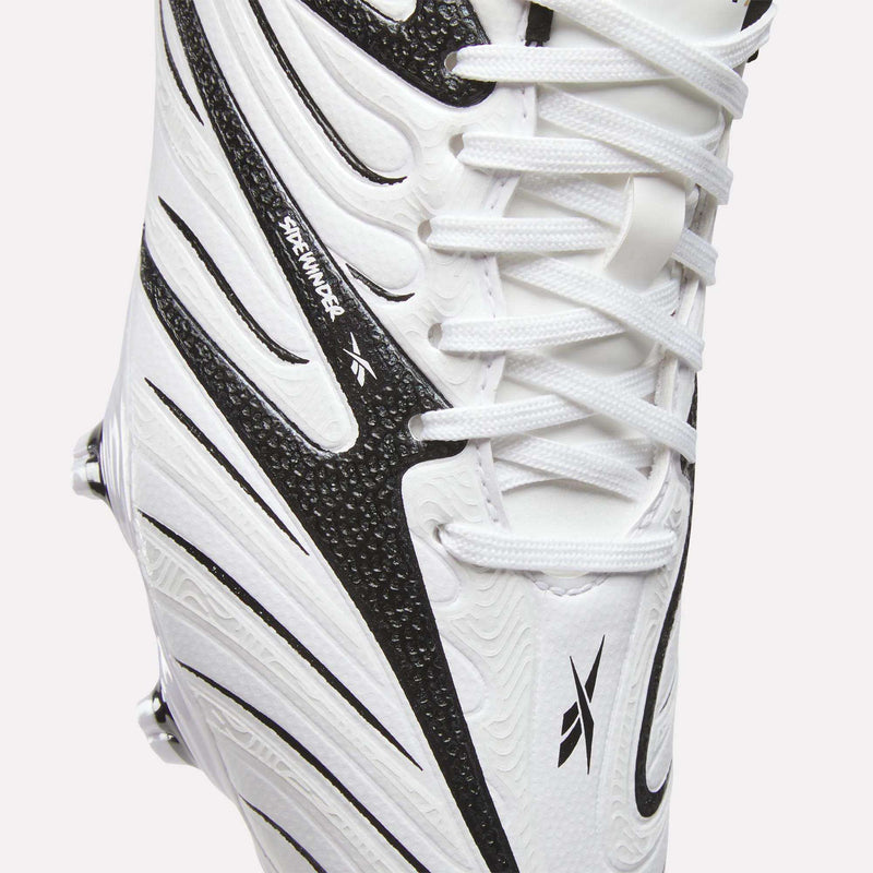 Close-up of the Reebok Sidewinder 26 League Firm Ground Cleats for Big Kids in white, featuring black textured details with dynamic black lines, visible laces and part of the sole. The design includes firm ground studs for optimal traction and a small Dreammaker label on the side.