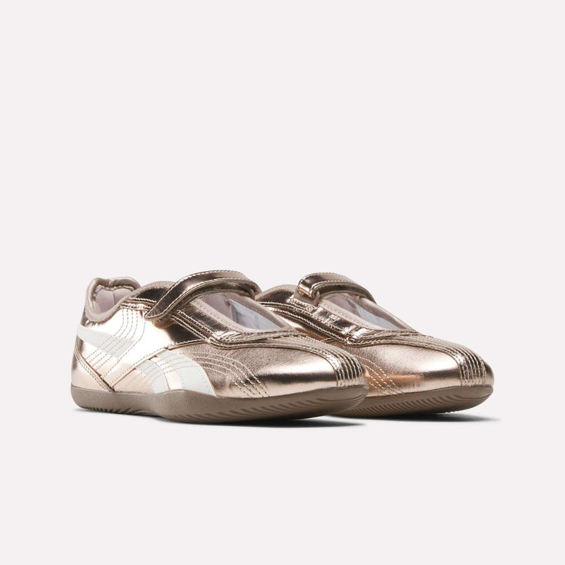 A pair of Reebok Women’s Ultra Lo Mary Jane Shoes in metallic rose gold leather, featuring white accents and Velcro straps, displayed on a plain light background.