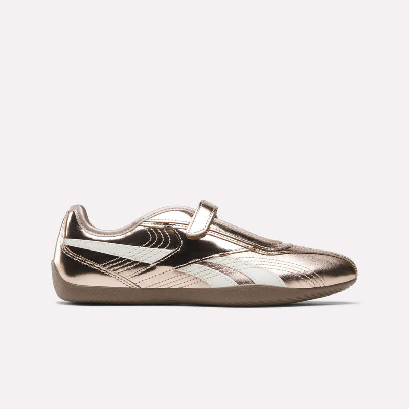 A metallic rose gold women’s sneaker with a white stripe detail, velcro strap, and flat sole is shown in a side profile against a plain white background—these Women’s Ultra Lo Mary Jane Shoes by Reebok combine elegant Rose Gold/Chalk leather accents for a contemporary update on classic style.