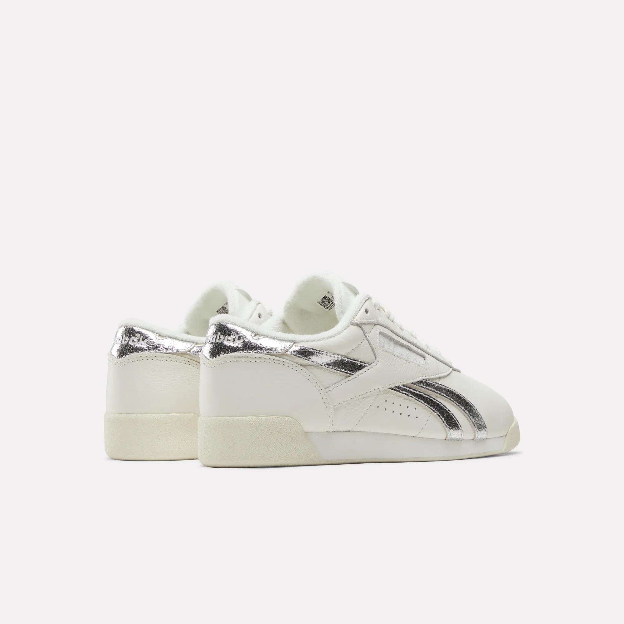 A pair of Reebok Women's Freestyle Lo Shoes featuring metallic accents on the heel and side stripes are displayed against a plain, light background with their heels angled slightly toward the viewer.