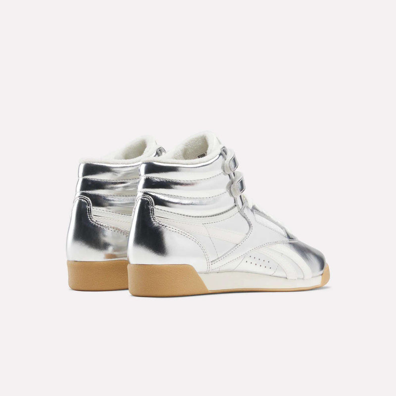 A pair of Women’s Freestyle Hi Shoes by Reebok, crafted from premium shiny silver leather with white midsoles and light brown rubber outsoles, displayed from an angled rear view against a plain white background—an elevated choice in women’s footwear.