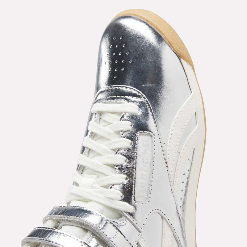 A close-up of the Reebok Women’s Freestyle Hi Shoes showcases their premium metallic silver leather finish, featuring white laces, a velcro strap, gum sole, and perforated toe for a stylish and elevated look.
