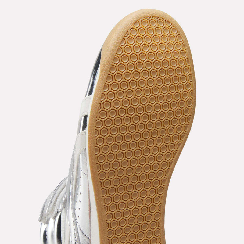 Close-up of the bottom of the Reebok Women’s Freestyle Hi Shoes, showcasing a light brown rubber sole with a hexagonal pattern and a partially visible upper crafted from premium silver and white leather.