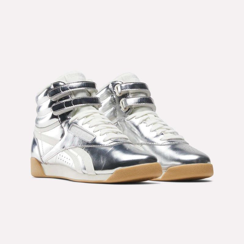 The Reebok Women’s Freestyle Hi Shoes are a pair of high-top sneakers crafted from premium leather, showcasing shiny silver and white panels, Velcro straps, white laces, and light brown rubber soles. These stylish women’s shoes are displayed on a plain light background.