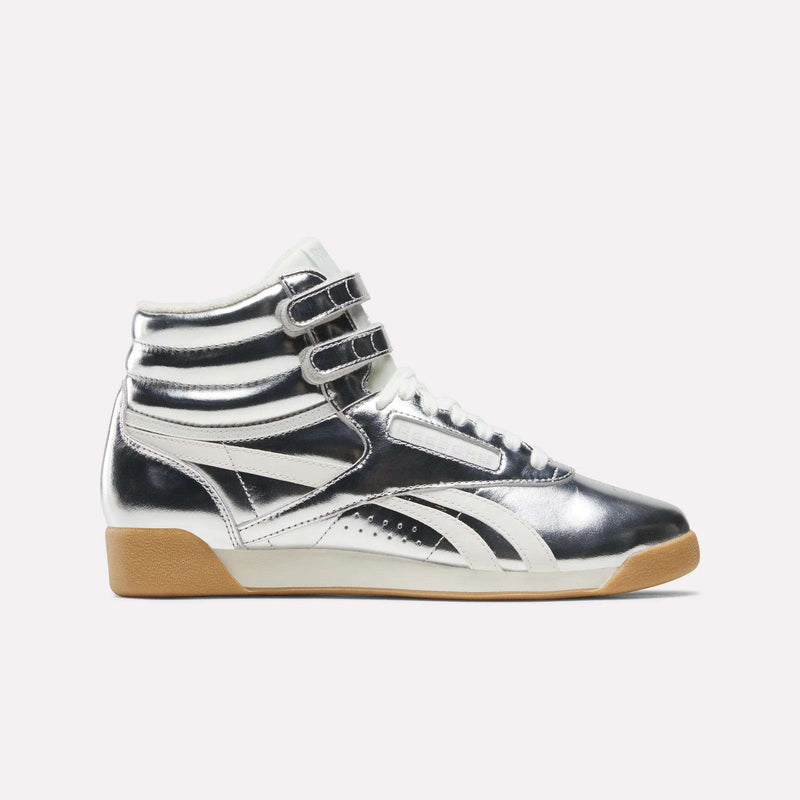 The Women’s Freestyle Hi Shoes by Reebok are high-top sneakers in a striking Silver Metallic/Chalk/Gum colorway, featuring premium leather construction, metallic silver finish with white accents, double velcro straps, white laces, and a tan rubber sole—perfectly capturing the bold style synonymous with Reebok—displayed against a plain white background.