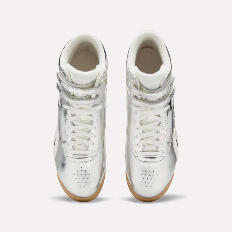 A pair of Women’s Freestyle Hi Shoes by Reebok in high-top, metallic silver premium leather, featuring Velcro straps and white laces, displayed from above on a plain light background.