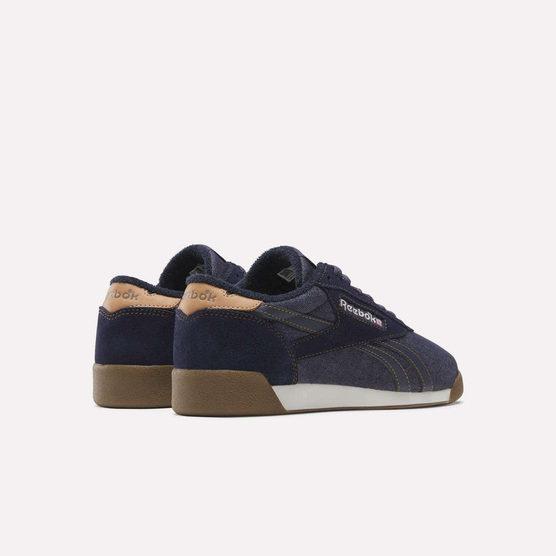 The Women’s Freestyle Lo Shoes by Reebok, featuring a navy blue design with tan accents on the heel and brown rubber soles, are displayed from the back and side against a white background, highlighting the classic charm of vintage-inspired denim sneakers for women.