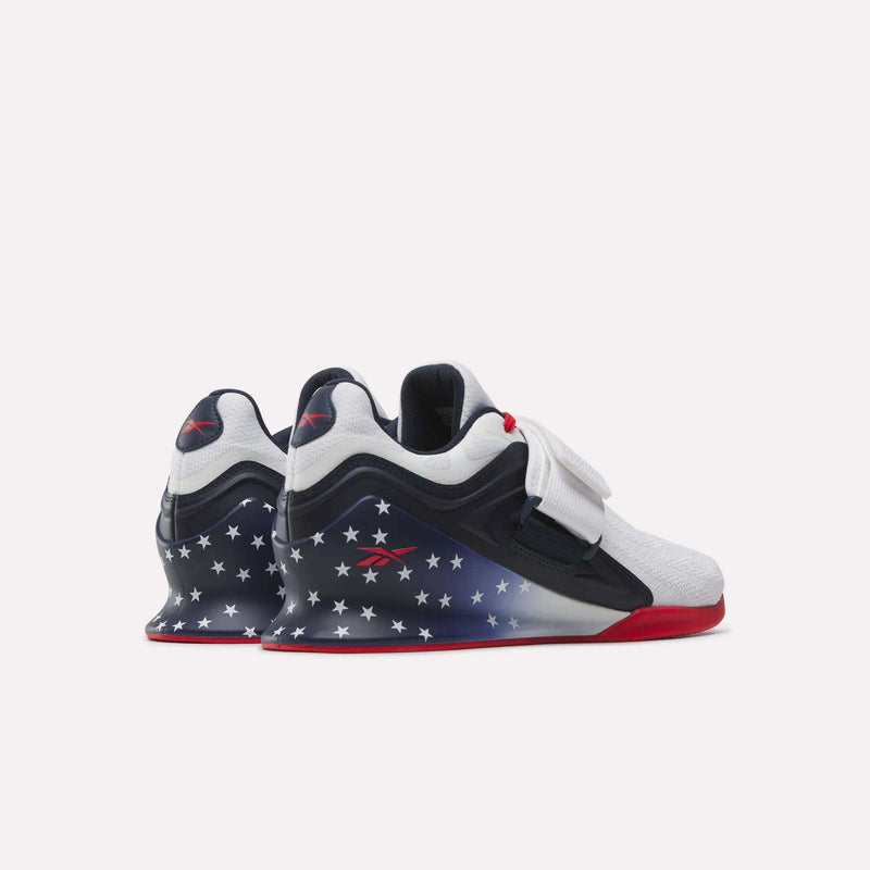 The Reebok Men's Legacy Lifter IV Shoes feature white uppers, navy blue sides with white stars, red soles, and white straps. Red Reebok logos on the heel and side add style and stability for your workouts.