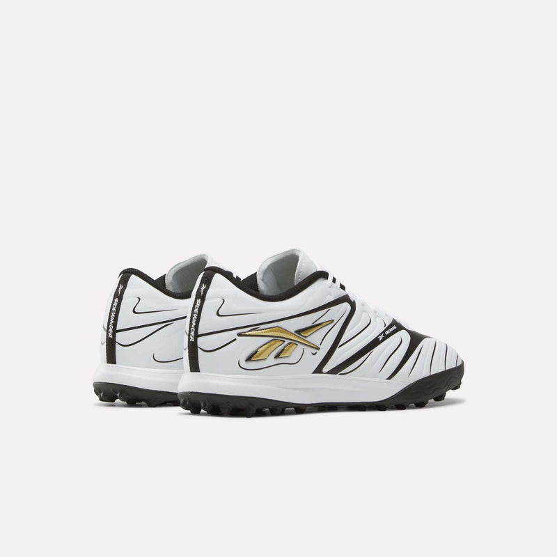 A pair of Reebok Sidewinder 26 League Turf Cleats in white with black accents, black soles, and a gold and black logo on the side, designed for turf players on artificial soccer fields, displayed against a plain light background.