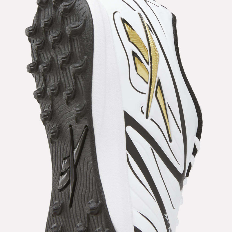 Close-up of the Reebok Sidewinder 26 League Turf Cleats in white, featuring black and gold accents. The shot highlights the textured black sole engineered for superior traction—ideal for turf players and delivering top performance on artificial soccer fields.