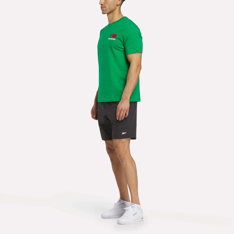 A man stands sideways wearing a green Reebok Mexico Football T-Shirt, black shorts with a small Reebok logo, and white sneakers against a plain white background.