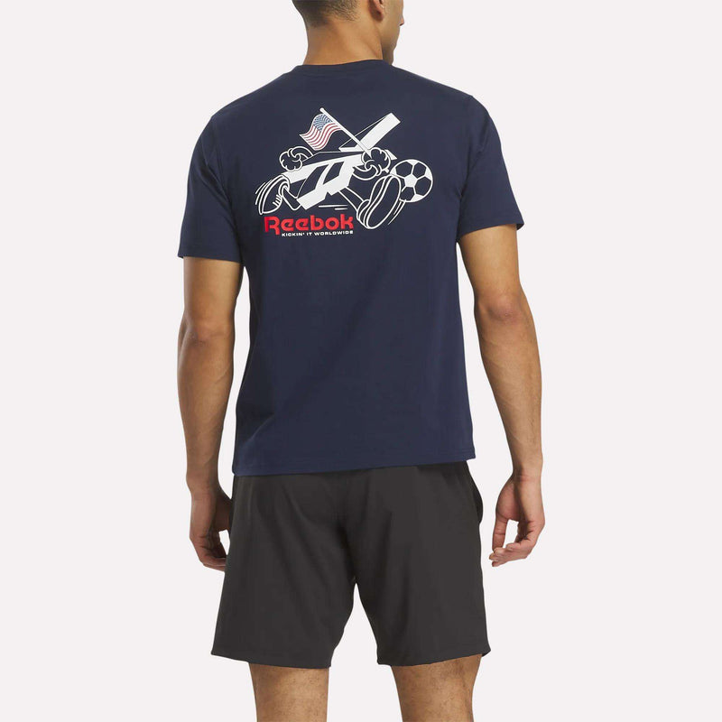 A man stands with his back to the camera, wearing a navy blue Reebok USA Football T-Shirt featuring sports equipment graphics and an American flag on the back, paired with black shorts.