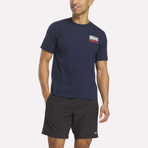 A man smiles in a navy blue Reebok USA Football T-Shirt, featuring soft cotton jersey and a small USA graphic on the left chest, paired with black athletic shorts. One hand is by his side, the other rests slightly in his pocket.