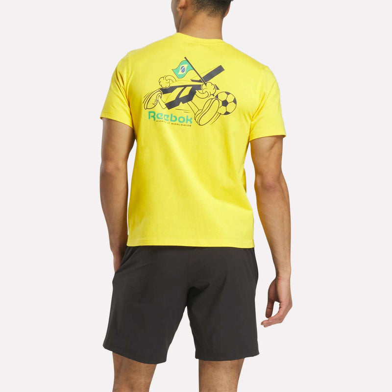 A person wearing a Reebok Brasil Football T-Shirt, featuring bright yellow cotton and a flag, soccer ball, and cartoon hands graphic on the back, is paired with black shorts and faces away from the camera.