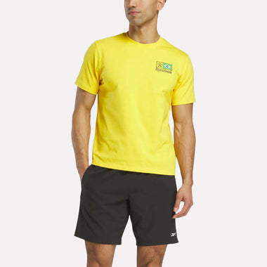 A person wears a bright yellow Reebok Brasil Football T-Shirt and black shorts, standing against a white background with arms relaxed and the Reebok logo on the t-shirt clearly visible.