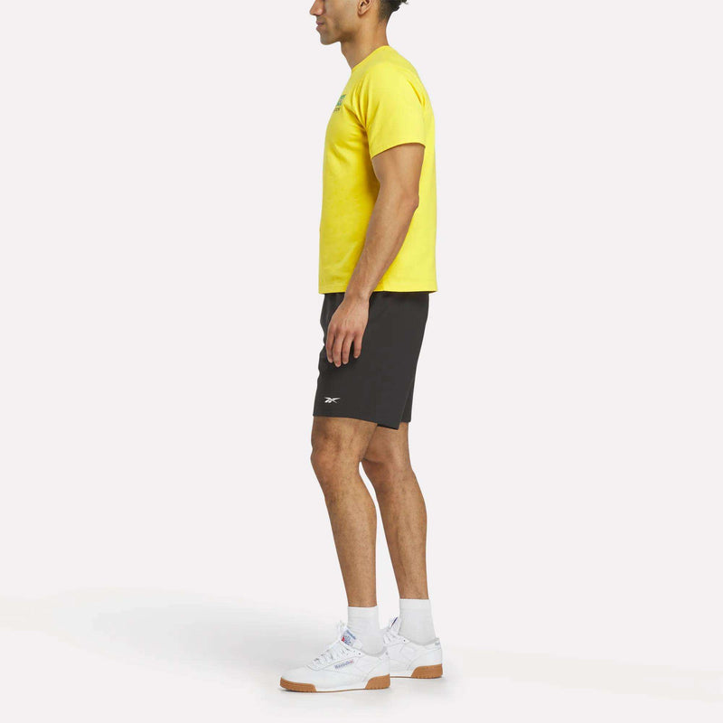 A man stands in profile wearing a yellow Reebok Brasil Football T-Shirt, black shorts, white socks, and white sneakers against a plain white background.